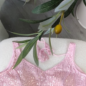 May include: A light pink sequined top with a V-neck and spaghetti straps. The top is embellished with numerous sequins, creating a shimmering effect. The top is displayed on a white surface, with green leaves and a yellow fruit in the background.