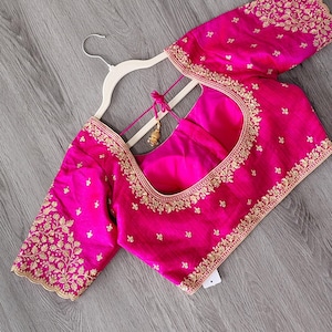May include: A vibrant fuchsia blouse with intricate gold embroidery along the neckline, sleeves, and edges. The blouse is displayed on a white hanger against a gray wooden surface. The sleeves are short and the neckline is a rounded square.