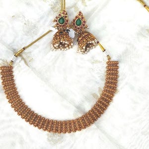 May include: A gold-toned necklace and matching earrings set. The necklace is a choker style with a series of connected beads. The earrings feature a green stone surrounded by red stones and a dangling bell shape with small white beads.