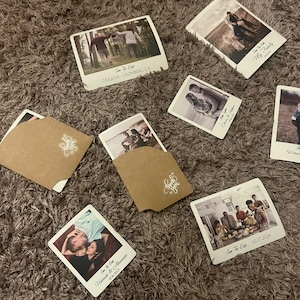K&ouml;nnte beinhalten: Ein Satz von 10 Save-the-Date-Karten im Polaroid-Stil. Die Karten zeigen Fotos eines Paares, einer Familie und einer Gruppe von Freunden. Die Karten sind auf wei&szlig;em Karton gedruckt und haben einen braunen Kraftpapierumschlag.