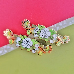 May include: Two gold-tone enamel pins featuring white flowers with green leaves and orange accents. The pins are in the shape of a floral sprig.