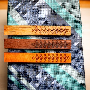 May include: Three wooden tie clips with a geometric pattern engraved on each. The tie clips are arranged vertically on a blue and white patterned fabric.