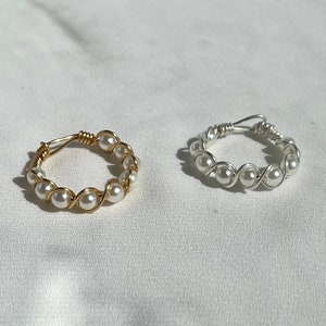 May include: Two handmade rings. One ring is gold-colored wire wrapped with white pearls. The other ring is silver-colored wire wrapped with white pearls. Both rings are on a white surface.