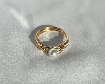 May include: A gold-coloured wire-wrapped ring with a single white pearl. The ring has a delicate design with the wire wrapped around the band and a pearl set in the centre. The ring is on a white surface.