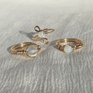 May include: Three gold-colored wire rings with a delicate design. Each ring features a small, round, opalescent stone. The rings are displayed on a white, textured surface, showcasing the jewelry's craftsmanship and style.