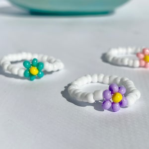 May include: Three white beaded rings with colorful flower charms. The rings have white beads and a single flower charm in turquoise, purple, and pink.