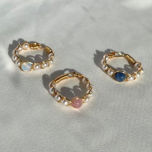 Könnte beinhalten: Drei goldene Ringe mit Perlenbesatz und jeweils einem Edelstein. Die Ringe haben einen hellblauen, rosafarbenen und dunkelblauen Edelstein.