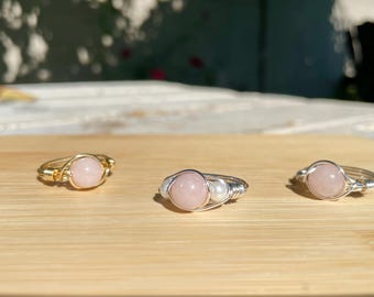 May include: Three wire-wrapped rings with rose quartz stones. One ring is gold-toned, while the other two are silver-toned. The rings are displayed on a light brown wooden surface, with sunlight in the background.