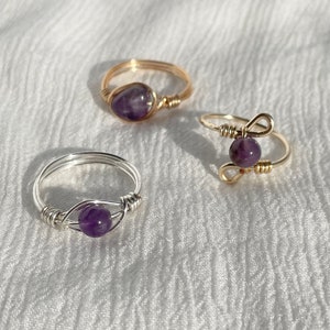 May include: Three wire-wrapped rings with purple amethyst stones. One ring is silver, and two are gold. The rings are displayed on a white, textured surface. The amethyst stones are round and vary in size.