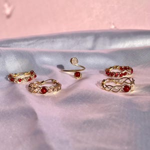 Può includere: Cinque anelli di filo d'oro con perline rosse e bianche. Gli anelli sono su uno sfondo di tessuto rosa.