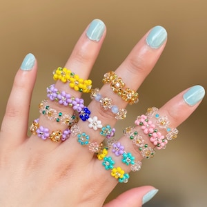 May include: A set of eight beaded rings featuring colorful flower designs. The rings are made with small, colorful beads and are arranged in a stack on a hand.