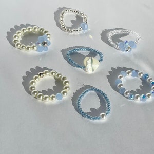 May include: A set of seven delicate rings featuring a variety of blue and white beads. Some rings have a single blue bead, while others have a cluster of blue beads. One ring has a white pearl and another has a white shell.