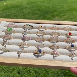 Op de afbeelding: Een display van handgemaakte draadgewikkelde ringen met diverse edelstenen en kralen. De ringen zijn gemaakt met goudkleurige draad en hebben een verscheidenheid aan kleurrijke stenen, waaronder roze, groene, blauwe en heldere edelstenen en parels. De ringen zijn gerangschikt op een lichtgekleurde tray.