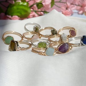 May include: A collection of ten gold-wrapped rings with different colored gemstones. The rings are arranged on a white fabric surface.