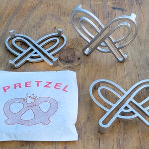 Può includere: Tre sottobicchieri in metallo argentato a forma di pretzel. I sottobicchieri hanno la forma di un pretzel, con due anelli collegati al centro. I sottobicchieri sono su una superficie di legno. La parola "PRETZEL" è stampata in rosso su un tovagliolo di carta bianco.