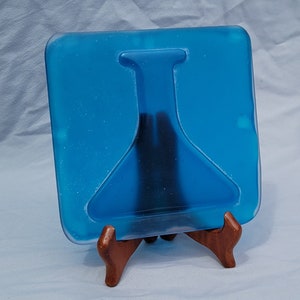 May include: A square, blue glass art piece with a dark, abstract design, displayed on a small wooden easel. The glass has a textured appearance and is a vibrant shade of blue.