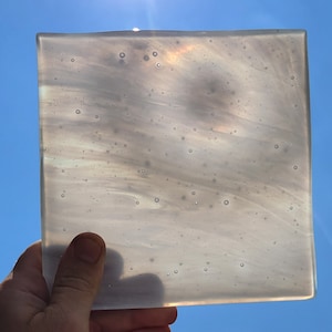Puede incluir: Una pieza de vidrio cuadrada y translúcida con un patrón nacarado y arremolinado. El vidrio tiene una superficie ligeramente texturizada y se sostiene contra un cielo azul brillante, con el sol brillando a través. Es probable que sea un artículo decorativo o un componente de una obra de arte más grande.
