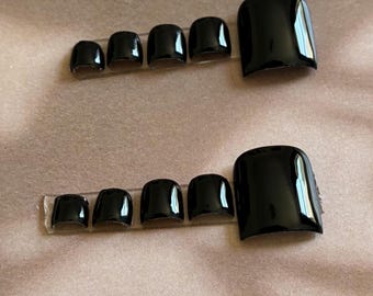 May include: Two sets of five glossy black press-on toenails arranged in descending size order on clear adhesive strips. The brown backing card features the white text See instruction on back at the top, indicating application details.