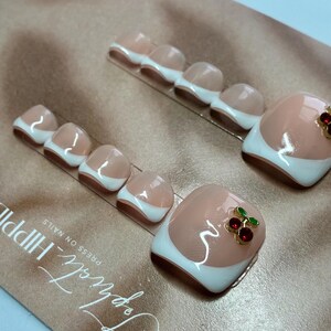 May include: Press-on toenails with a French tip design. The nails are a nude color with white tips. One nail has a cherry charm with red gems and green leaves. The nails are on a gold-colored surface.