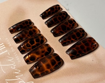 May include: A set of ten coffin-shaped press-on nails featuring a glossy brown and black tortoiseshell pattern. The nails are arranged in two rows on a light tan display card that includes the text Sophisti-Hills PRESS ON NAILS.