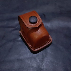 Könnte beinhalten: Ein braunes Lederetui mit einem schwarzen Knopfverschluss. Das Etui ist für die Aufbewahrung eines kleinen Gegenstands wie eines Feuerzeugs oder einer kleinen Taschenlampe gedacht.