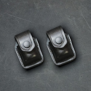 Peut inclure: Deux étuis en cuir noir avec une fermeture à bouton. Les étuis sont sur une surface texturée grise.
