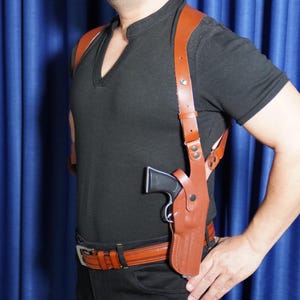 May include: A tan leather shoulder holster system featuring adjustable straps and a metal buckle, designed to carry a handgun. The holster is worn over a black shirt and includes a matching tan leather belt with a silver-toned metal buckle.