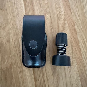 May include: A black leather pouch with a snap button closure and a separate black cylindrical component featuring a ribbed top and an exposed silver metal spring mechanism, both displayed together on a light-coloured wooden surface.