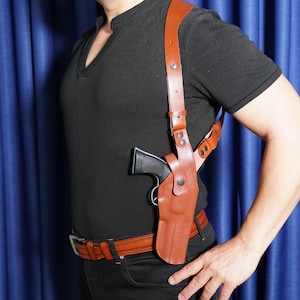 May include: A tan leather shoulder holster system featuring adjustable straps with metal hardware and a snap closure, designed to carry a black handgun. The holster is worn over a black short-sleeved shirt and paired with a matching tan leather belt.