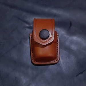 May include: A brown leather case with a black button closure. The case is designed to hold a small object, such as a lighter or a keychain.