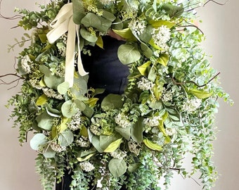 May include: A large, lush green wreath with white flowers and a white satin ribbon. The wreath is made of eucalyptus and other greenery.