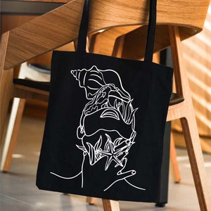 May include: A black tote bag with a white line drawing of a seashell, starfish, and seaweed. The bag is hanging on a wooden chair.