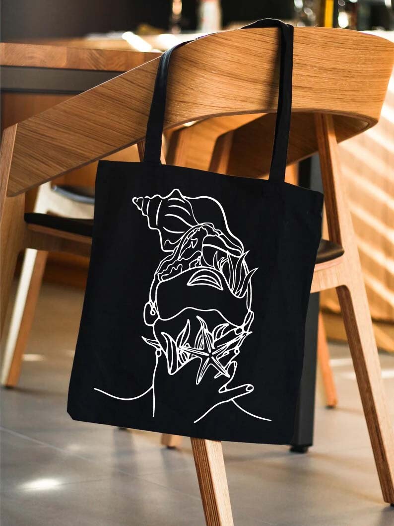 May include: A black tote bag with a white line drawing of a seashell, starfish, and seaweed. The bag is hanging on a wooden chair.