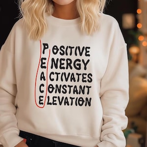 May include: Cream-colored sweatshirt with the words "POSITIVE ENERGY ACTIVATES CONSTANT ELEVATION" in black, with the word "PEACE" highlighted in red. The sweatshirt is a casual, comfortable style.