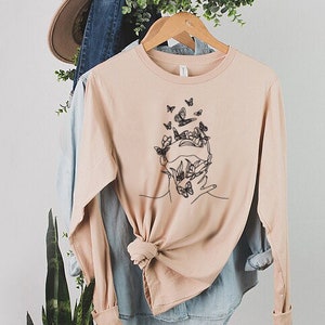 May include: A light pink long-sleeved t-shirt with a graphic of a face surrounded by butterflies and flowers. The shirt is made of a soft, comfortable material.
