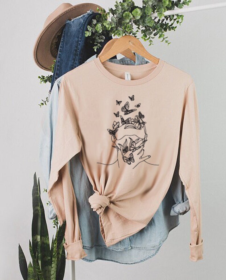 May include: A light pink long-sleeved t-shirt with a graphic of a face surrounded by butterflies and flowers. The shirt is made of a soft, comfortable material.