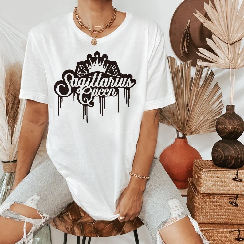 以下が含まれることがあります： 黒いグラフィックデザインが施された白いTシャツ。テキストの上に王冠とダイヤモンドが描かれており、「射手座の女王」と書いてあります。
