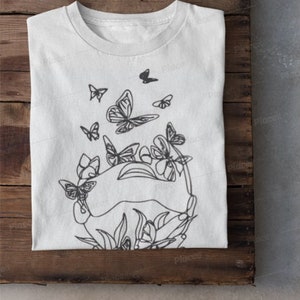 May include: A white t-shirt with a black line drawing of butterflies and flowers. The design features a circle of butterflies surrounding a cluster of flowers.