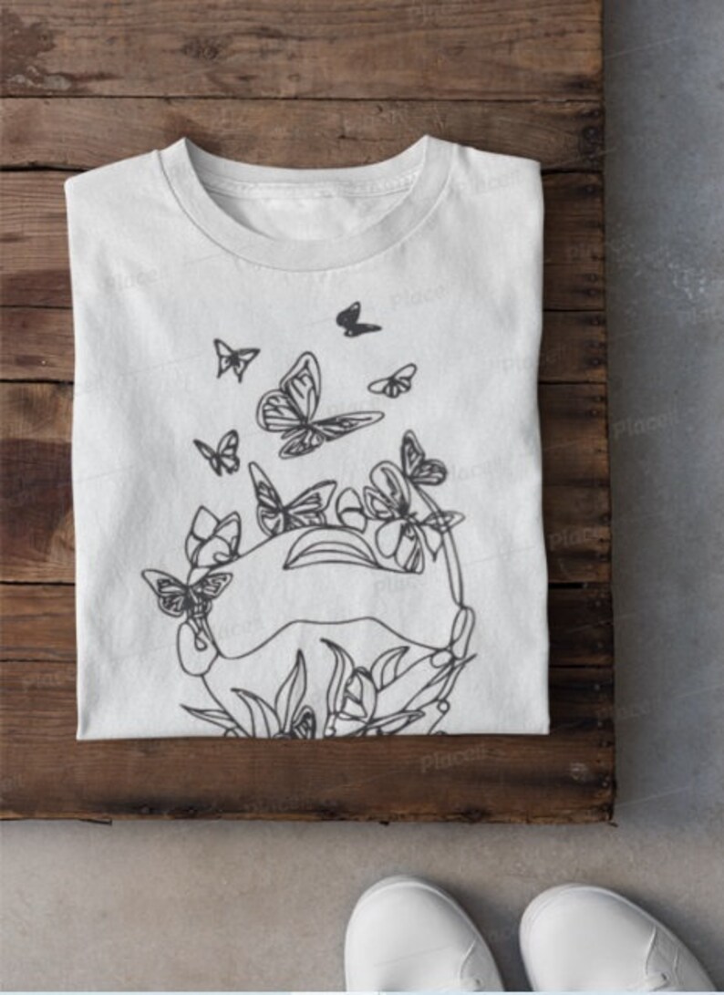 May include: A white t-shirt with a black line drawing of butterflies and flowers. The design features a circle of butterflies surrounding a cluster of flowers.
