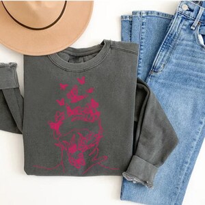 May include: A dark gray sweatshirt with a pink graphic of a face surrounded by butterflies. The sweatshirt is laid out on a white surface with a brown hat and blue jeans.