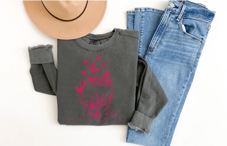 May include: A dark gray sweatshirt with a pink graphic of a face surrounded by butterflies. The sweatshirt is laid out on a white surface with a brown hat and blue jeans.