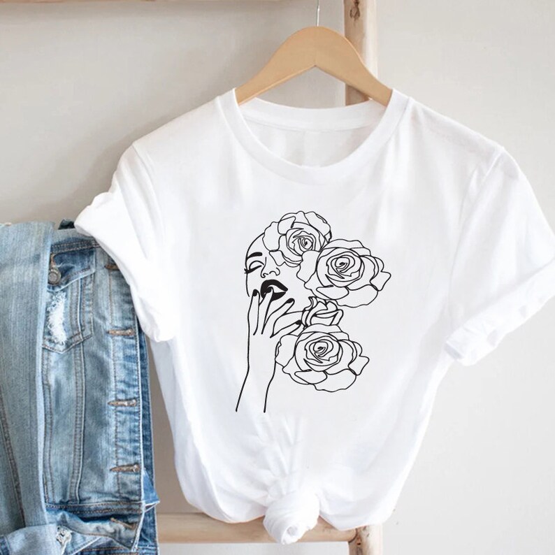 May include: White t-shirt with a black line drawing of a woman's face with roses around her head.