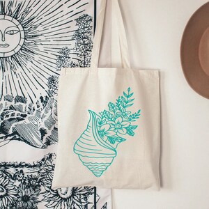 May include: A white canvas tote bag with a teal blue seashell design. The seashell is filled with teal blue flowers and leaves.