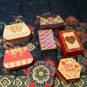 May include: An assortment of decorative gift boxes in various shapes and sizes. The boxes feature floral, geometric, and heart designs, with colors including pink, red, blue, and white. Some boxes have handles or the text "BEE HAPPY" and "BIG".