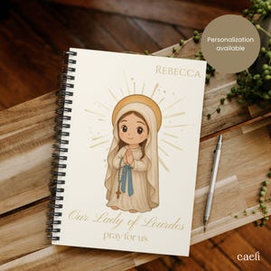 May include: Spiral-bound notebook featuring an illustration of Our Lady of Lourdes. The artwork depicts a cartoon-style figure with a halo, praying hands, and a rosary. The notebook has the name "Rebecca" printed at the top and the text "Our Lady of Lourdes pray for us" at the bottom.