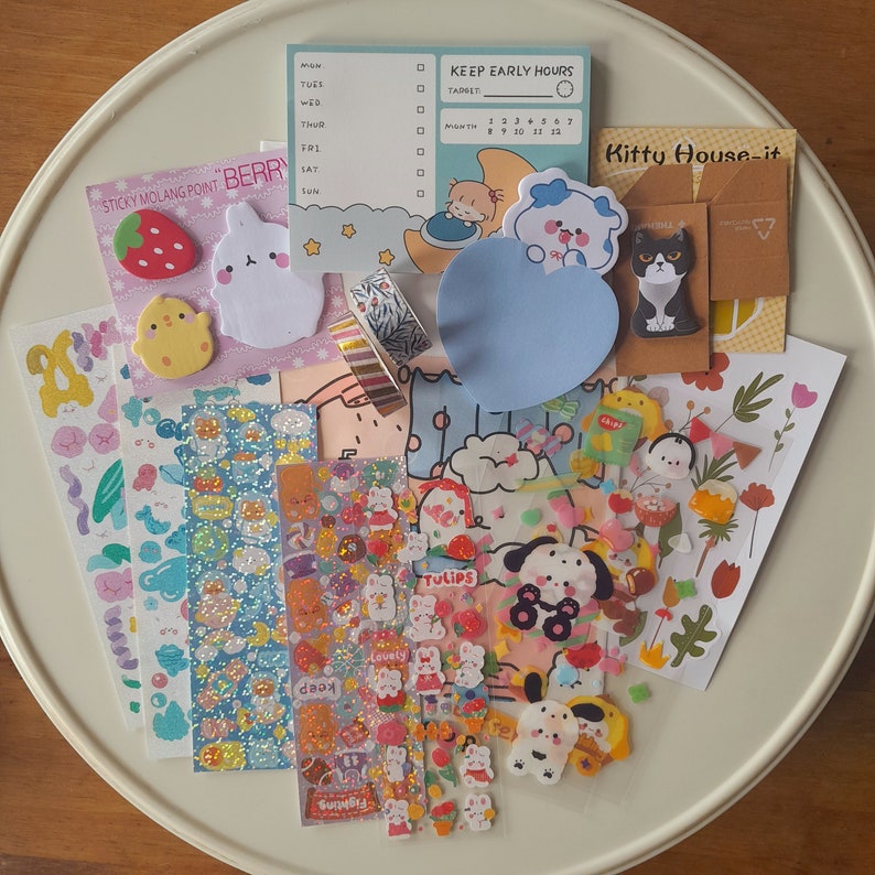 Puede incluir: Una colecci&oacute;n de pegatinas y papeler&iacute;a coloridas con lindos animales de dibujos animados, flores y otros dise&ntilde;os. Las pegatinas est&aacute;n dispuestas en un plato blanco con un fondo de madera clara. El texto "Kitty House-it" es visible en una pegatina marr&oacute;n.