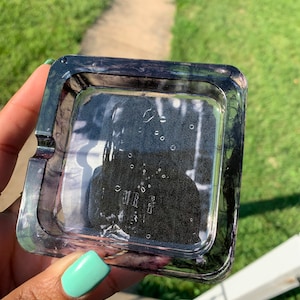 May include: A square glass ashtray with a black and white marbled design. The ashtray has a raised edge and a smooth surface.