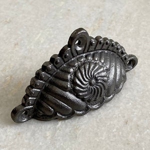Puede incluir: Un tirador de cajón de hierro fundido con un diseño decorativo de espiral. El tirador es de color gris oscuro y tiene un acabado ligeramente envejecido.