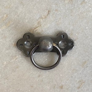 May include: A black metal ring pull handle with a decorative design. The handle is attached to a metal plate with two holes for screws.