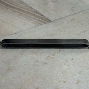 May include: A black metal drawer pull with a rectangular shape and rounded edges. The pull has two small, round holes on each end for mounting.